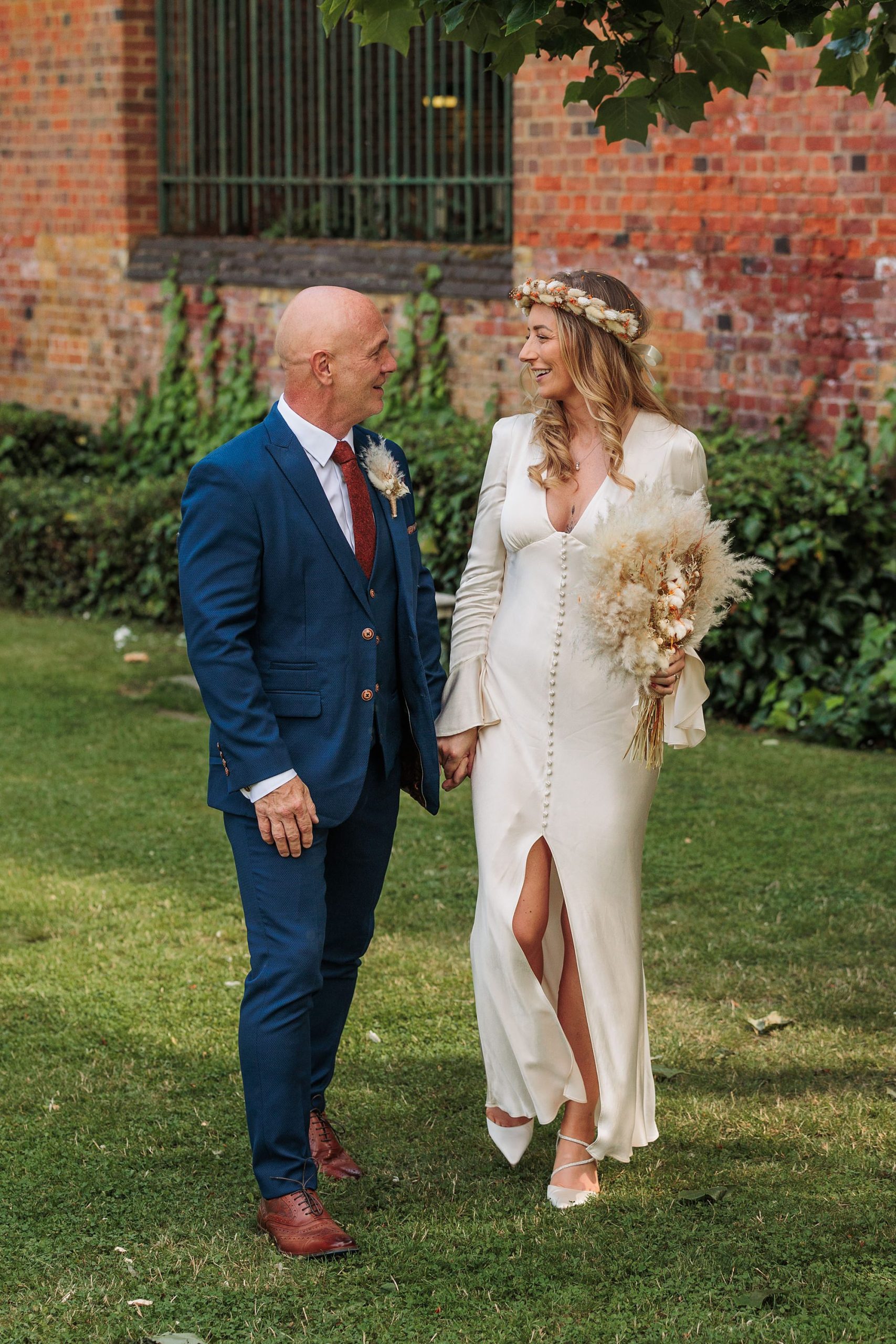 A couple stands on the grass in front of a brick wall at St Albans Registry Office. The woman, in a white gown, holds a bouquet of dried flowers. The man dons a blue suit with a brown tie. They smile at each other beneath the trees lush green leaves.