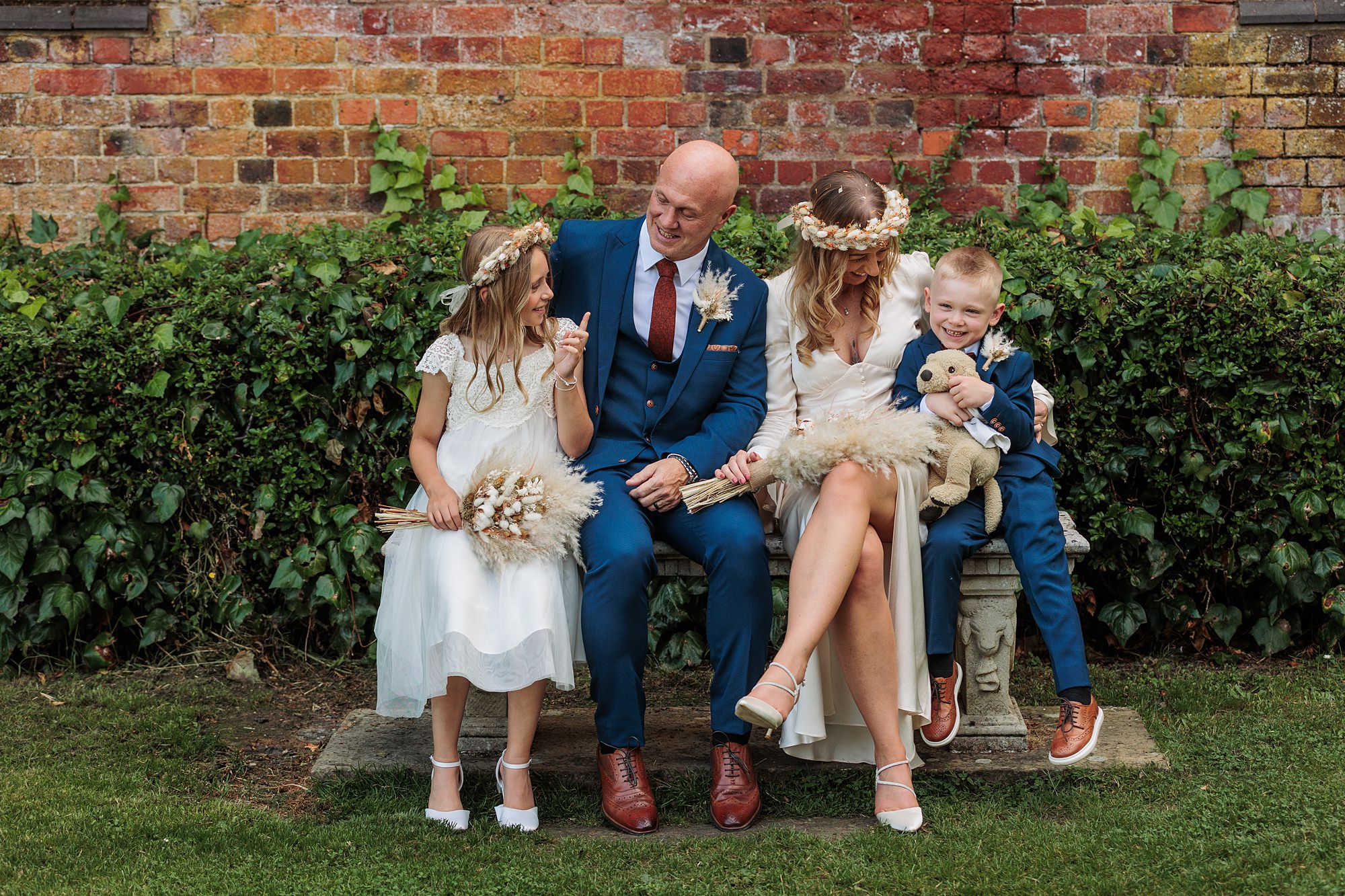A family of four, dressed in formal attire, sits on a bench in front of an ivy-covered brick wall at St Albans Registry Office. The parents, smiling, hold hands with their children. The girl wears a flower crown, and the boy holds a teddy bear.