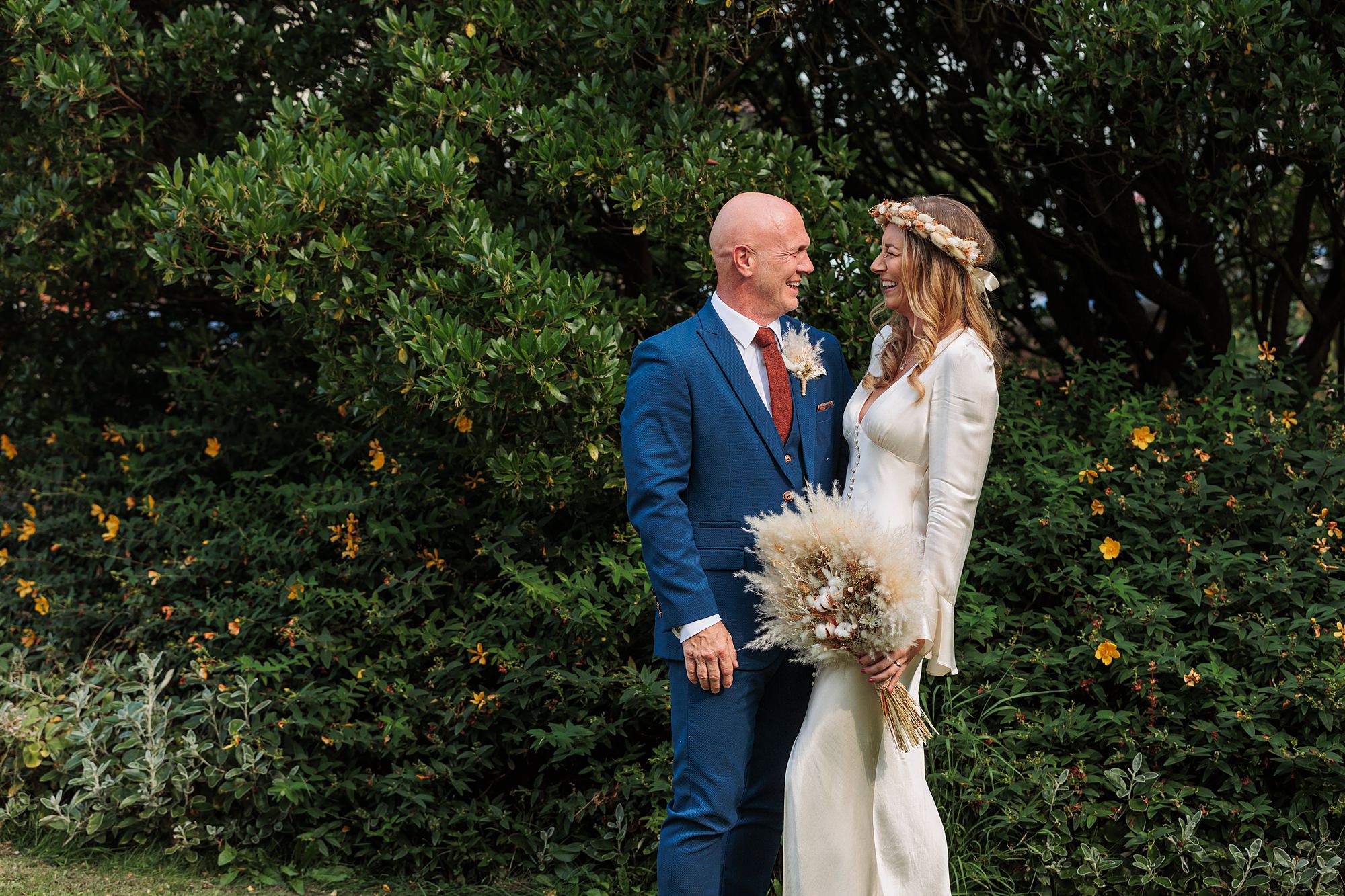 In the charming gardens outside St Albans Registry office, a couple stands smiling at each other. The person on the left wears a blue suit, while the one on the right is in a white dress with a floral headpiece, holding a bouquet amidst greenery and small yellow flowers.