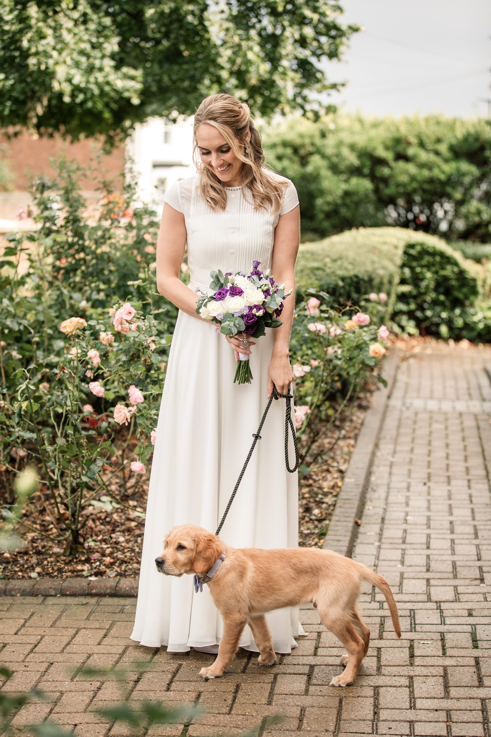 A smiling bride in a white dress holds a bouquet of flowers while standing on a paved walkway outside the St Albans Registry Office. She is gently holding the leash of a light brown puppy. Roses and shrubs surround the path, with trees visible in the background.