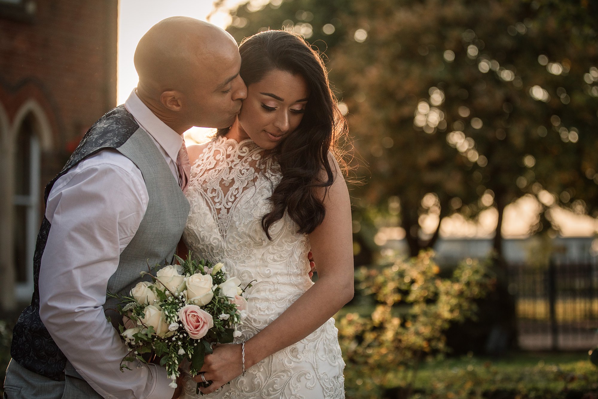 A groom gently kisses the brides head as they stand together in the golden sunlight outside St Albans Registry Office. The bride holds a bouquet of white and pink roses, framed by a blurred garden backdrop.