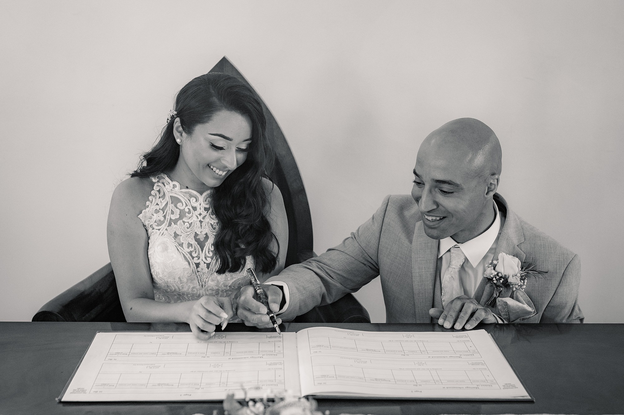 A bride in a lace dress and a groom in a suit are sitting at a table, smiling as they sign a document together. The scene at St Albans Registry office is both joyful and formal.