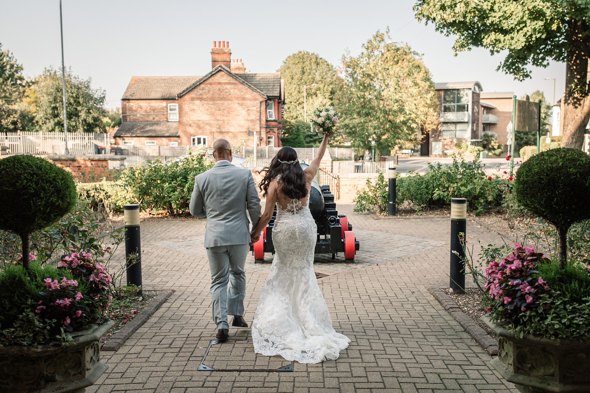 The bride in a white lace gown and groom in a gray suit walk hand in hand down the brick pathway of St Albans Registry Office, holding a bouquet aloft. The scene is outdoors with lush greenery and the elegant brick building as their backdrop.