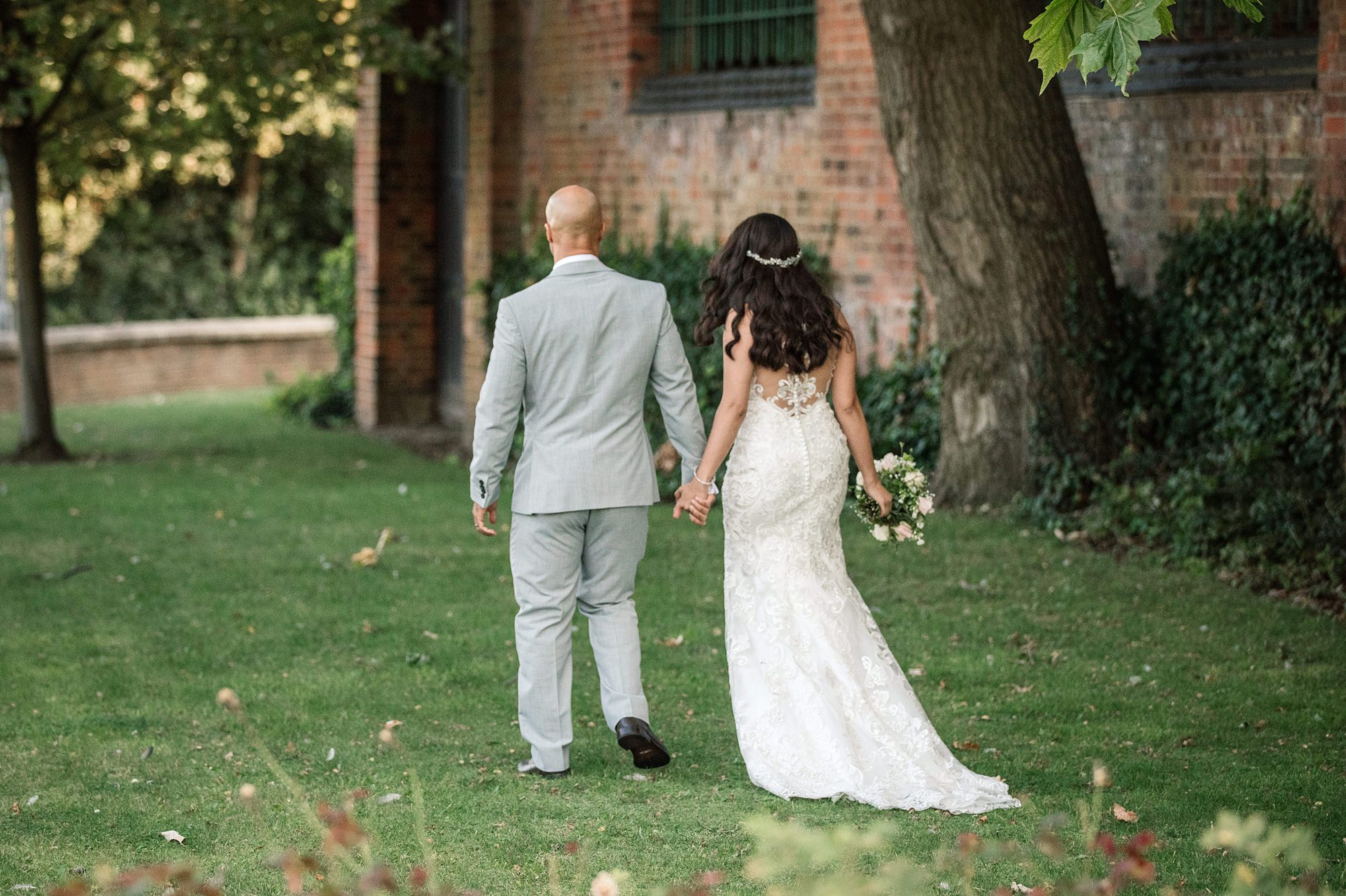 A bride and groom walk hand in hand on a grassy path beside the brick facade of St Albans Registry Office. The groom wears a light gray suit, and the bride shines in her white lace gown, clutching a small bouquet. Green trees and leaves frame this picturesque scene.