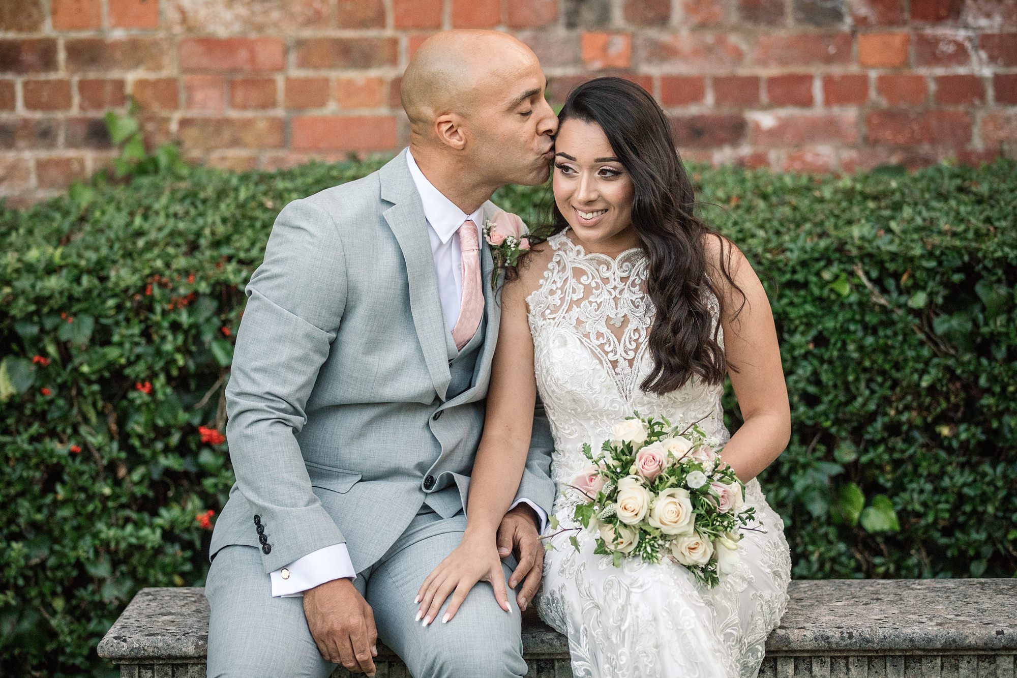 A bride in a white lace dress holds a bouquet of roses while sitting on a stone bench. Beside her, the groom in a light gray suit tenderly kisses her forehead. They share this romantic moment at the charming St Albans Registry Office, framed by a green hedge and brick wall.