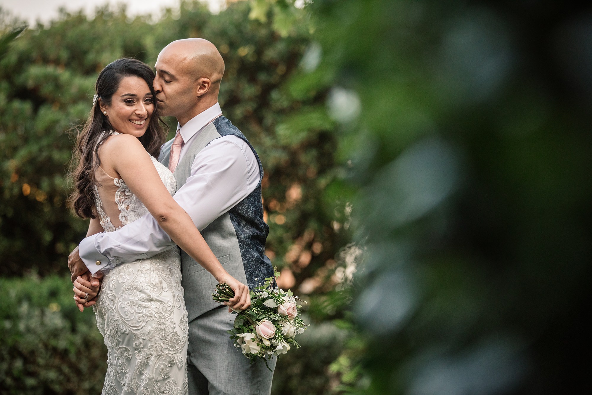 In a lush garden setting near St Albans Registry Office, a couple embraces lovingly. The groom kisses the bride on the temple as she smiles warmly at the camera. She holds a small bouquet, and both are dressed in elegant wedding attire.