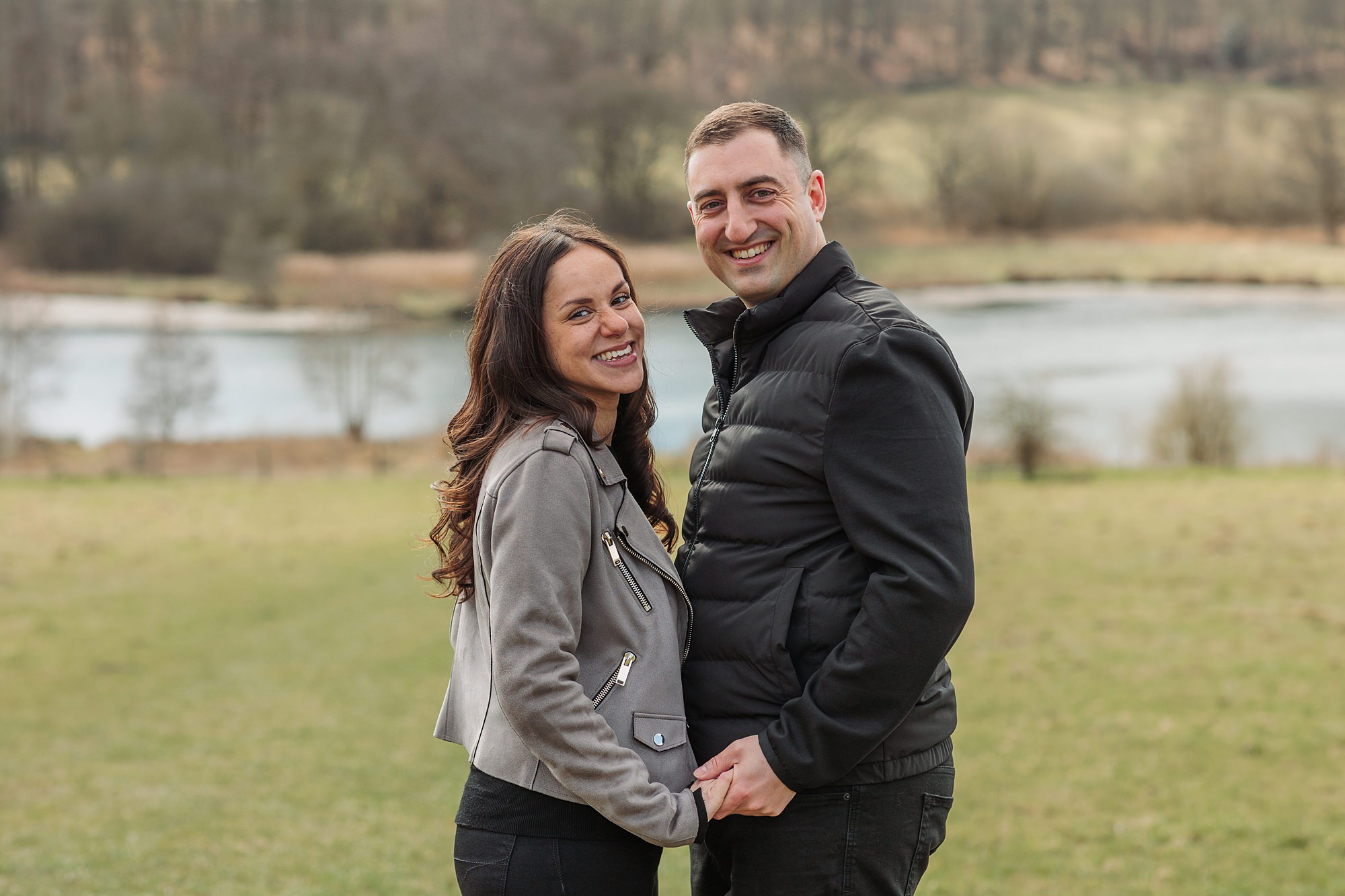 A smiling couple stands in a grassy outdoor setting, holding hands. The woman wears a gray jacket, and the man wears a black jacket. A lake and trees are visible in the blurred background.