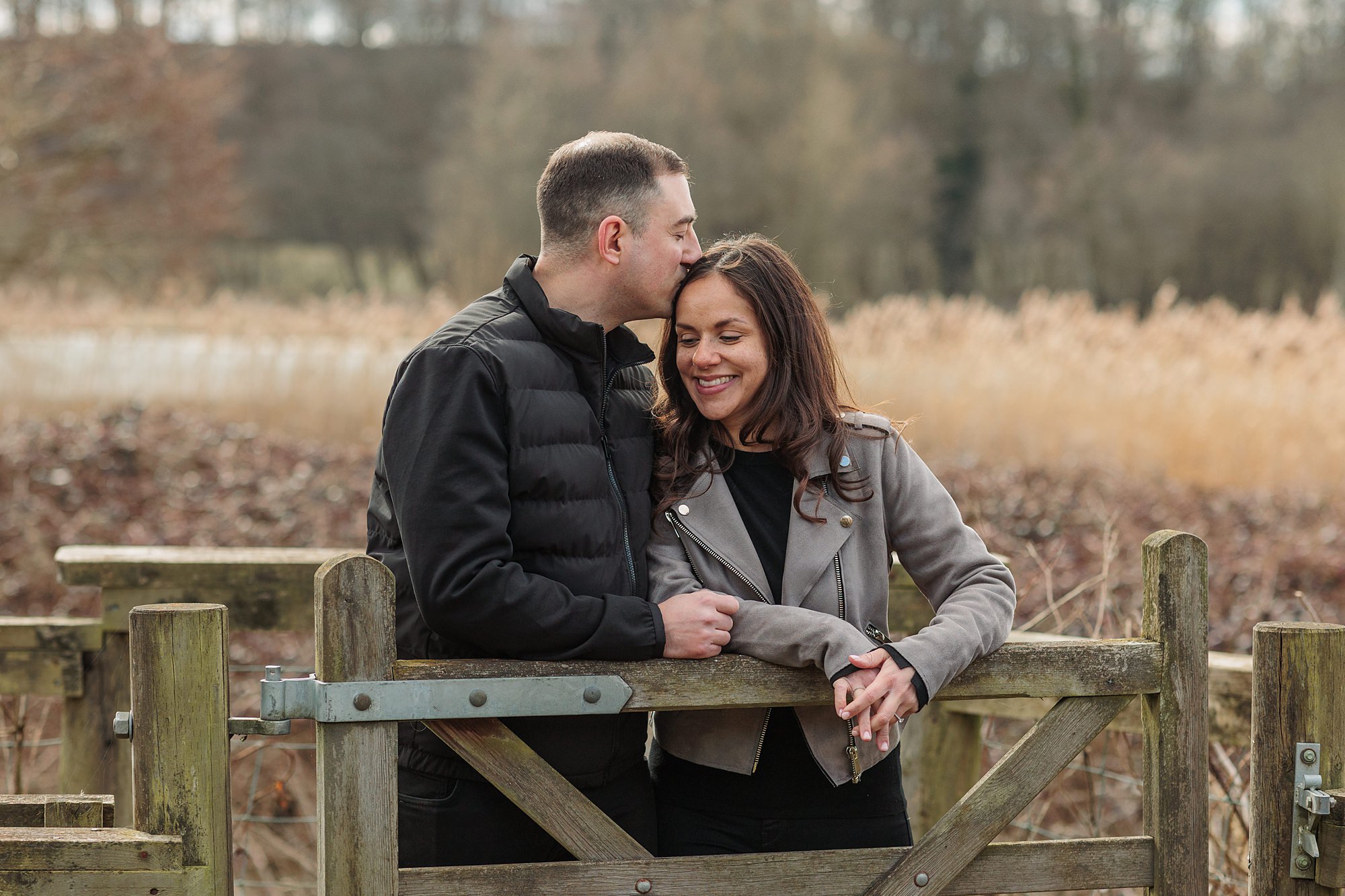 A man and woman stand together outside, leaning on a wooden gate. The man in a black jacket kisses the womans head. The woman, in a gray jacket, smiles with eyes closed. Dry grass and trees are blurred in the background.