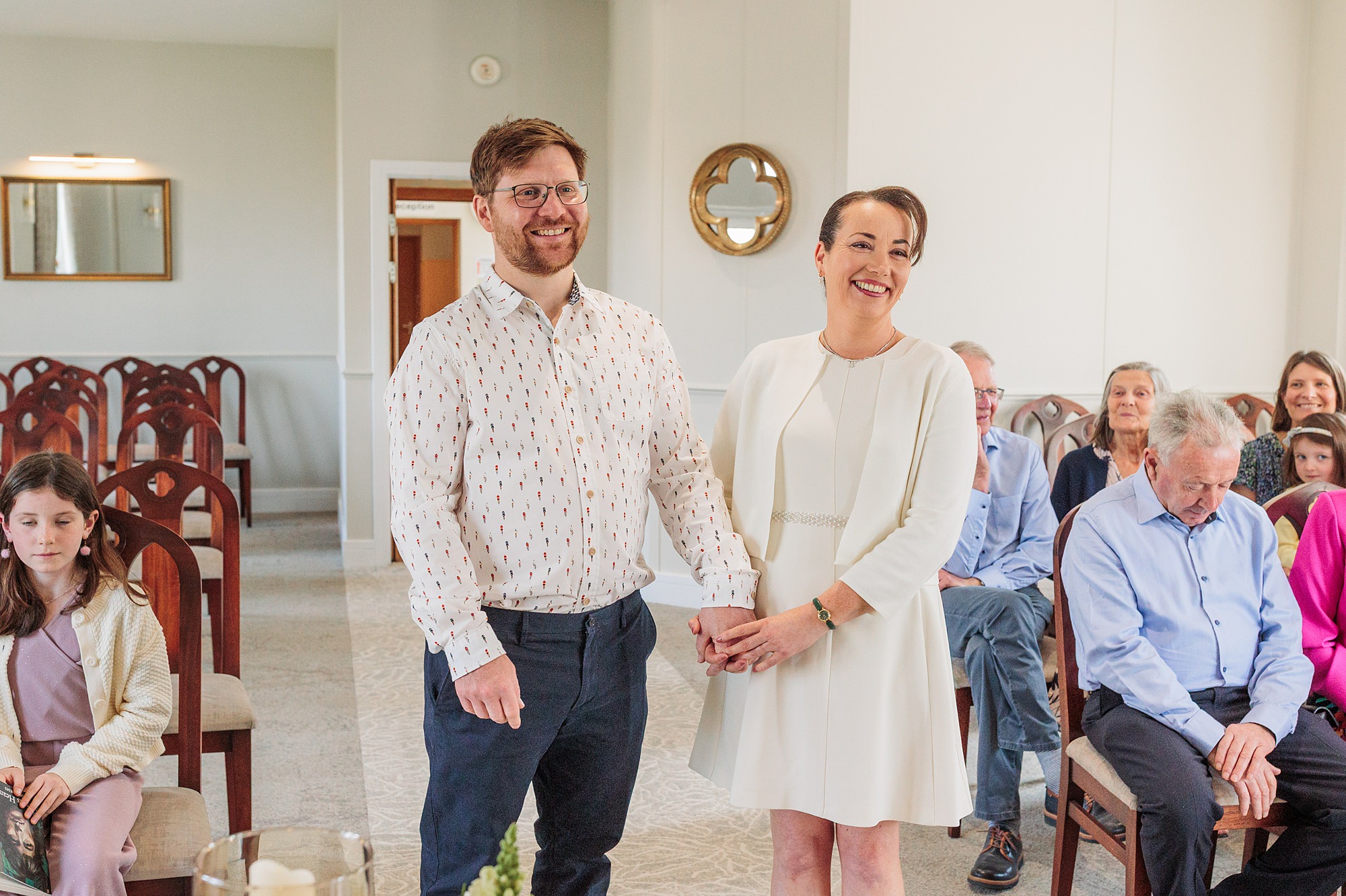 A man and a woman stand at the front of a room, holding hands and smiling, while seated guests watch. The room has white walls, wooden chairs, and natural light coming in.
