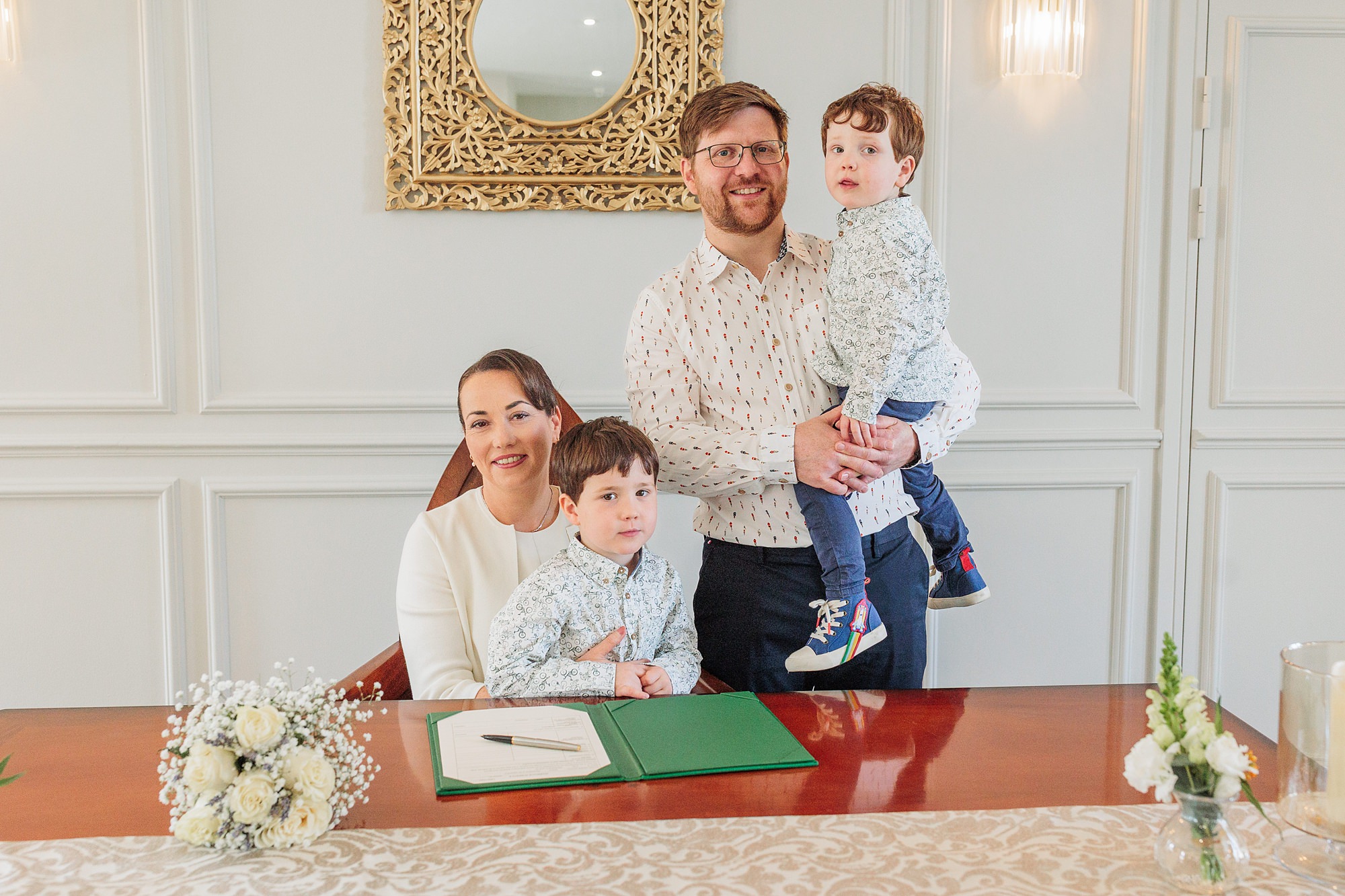A woman is seated at a table with a pen and open folder, and a man stands beside her holding a young boy. Another boy sits at the table. Flowers are on the table; a mirror hangs on the wall behind them.