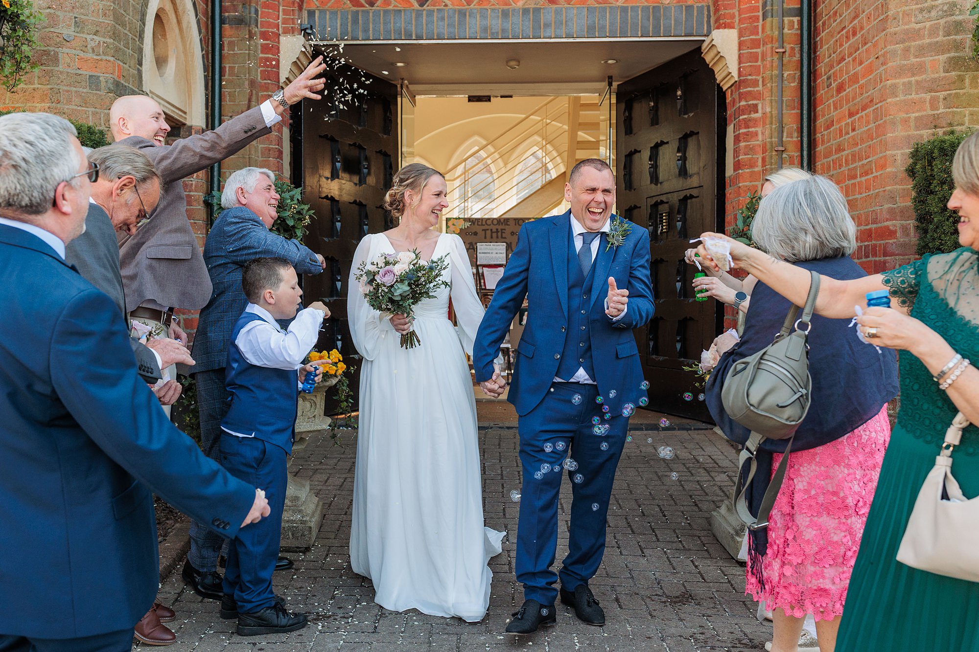 Newlywed couple exits a building holding hands, smiling, as guests on either side celebrate by throwing confetti and blowing bubbles. The bride wears a white dress and holds a bouquet; the groom wears a blue suit.