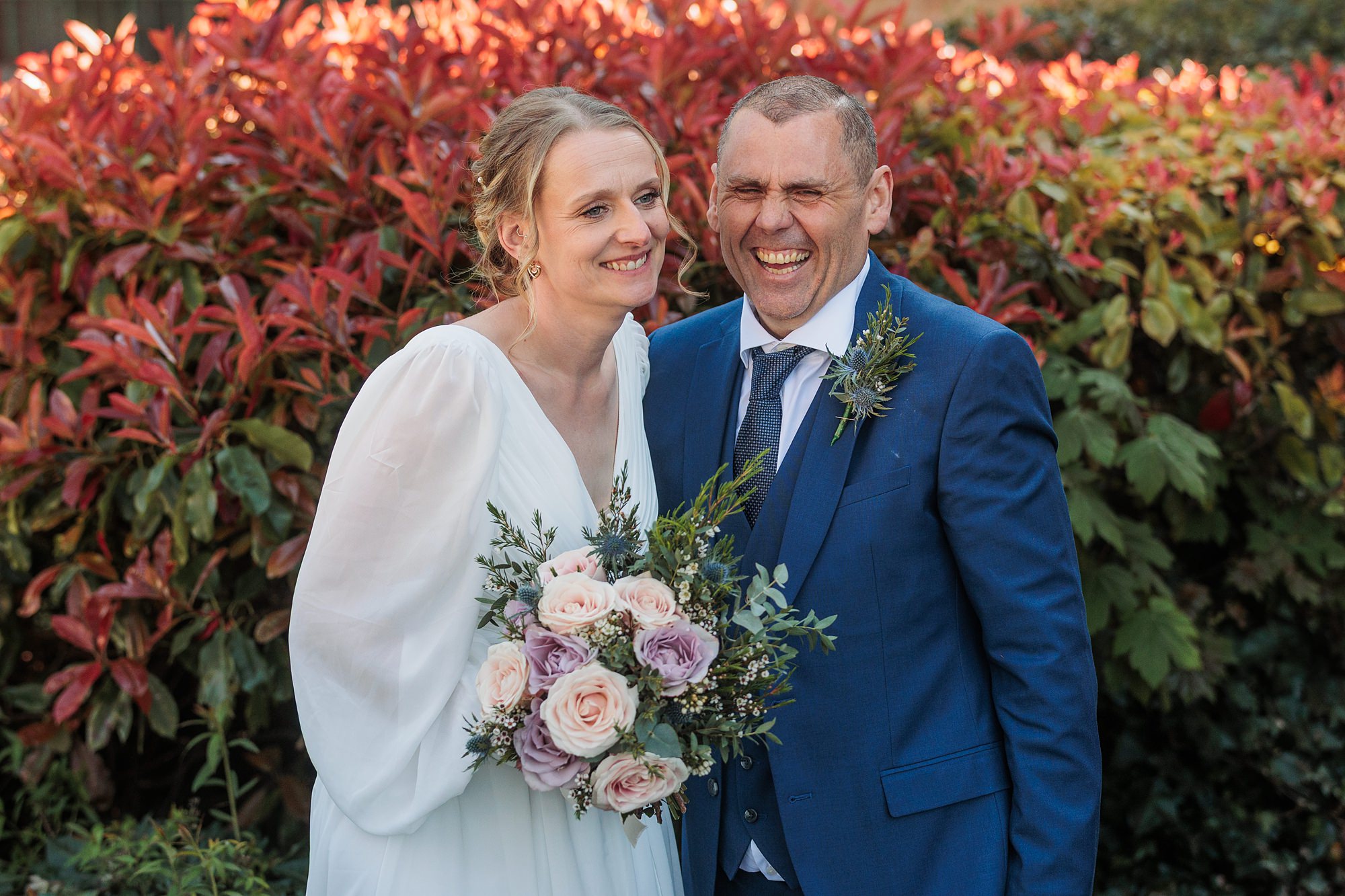 A bride in a white dress holds a bouquet of pale roses while standing next to a man in a blue suit and tie. They are smiling in front of a bush with red and green leaves.