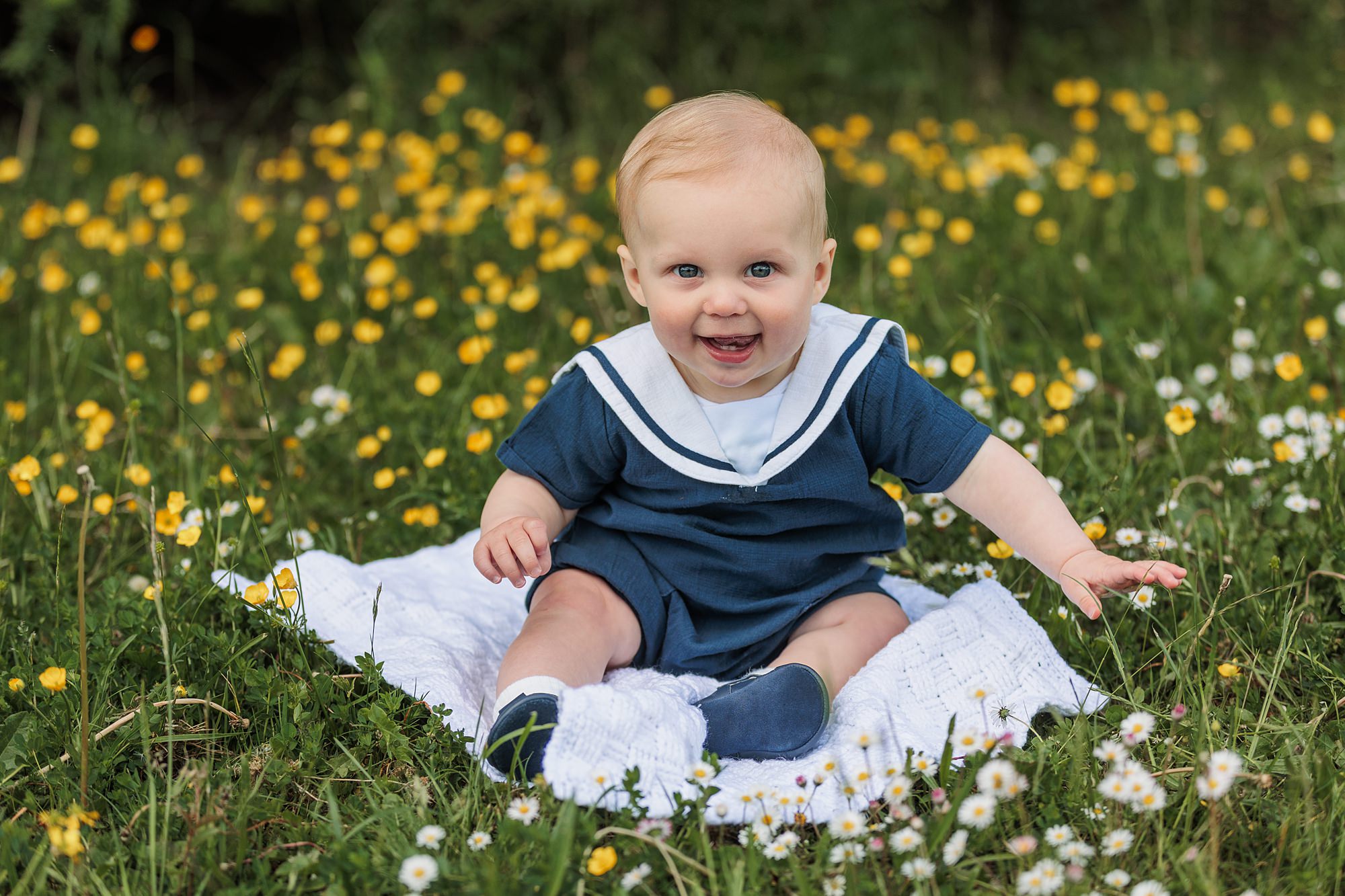A baby with blond hair sits on a white blanket in a field of grass and yellow and white flowers, wearing a blue outfit with a white sailor-style collar, and looks forward smiling.
