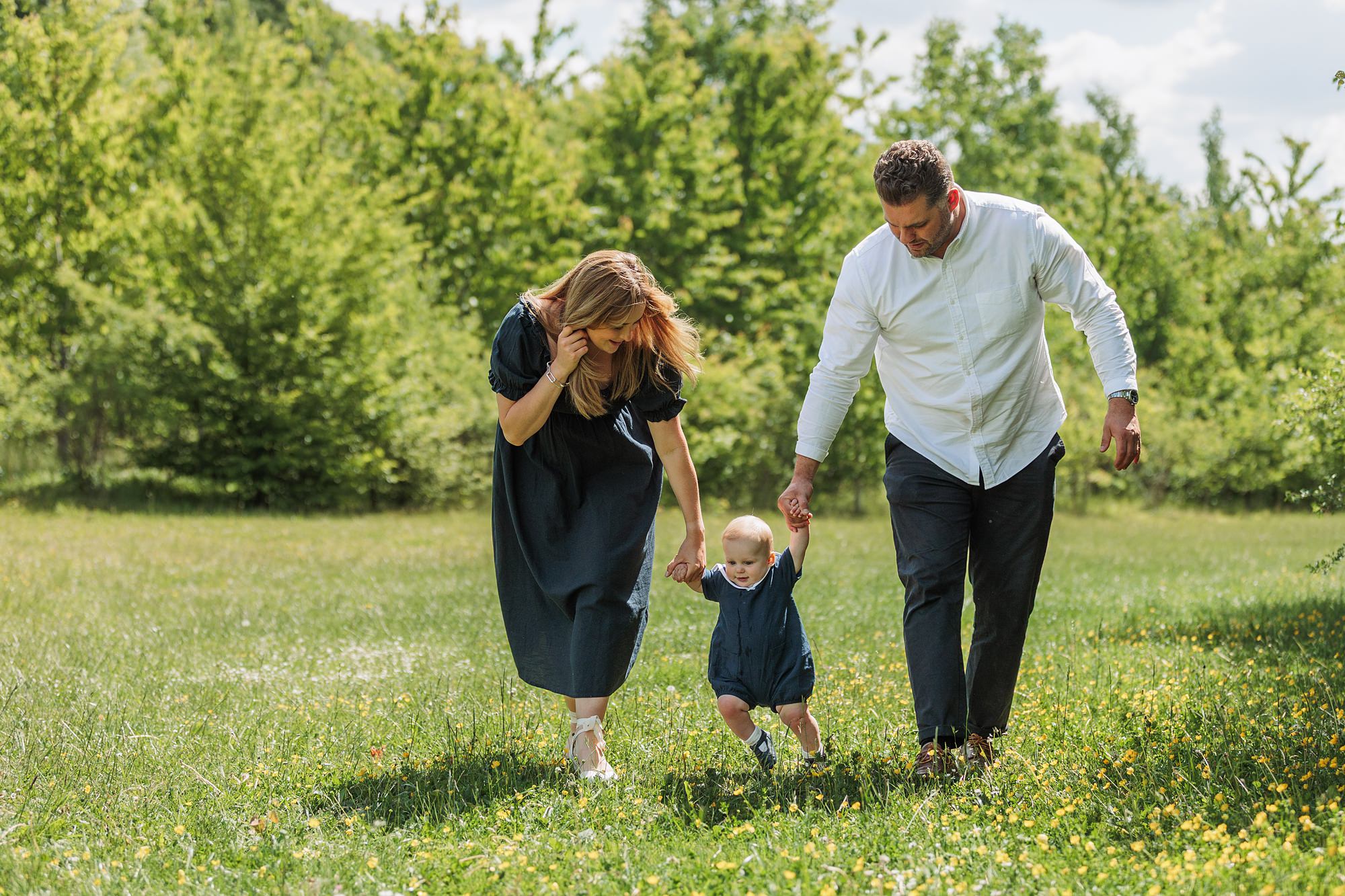 A woman and a man hold hands with a small child as they walk together in a grassy, sunny field surrounded by trees. The adults are dressed in dark and light clothing, and the child wears a blue outfit.