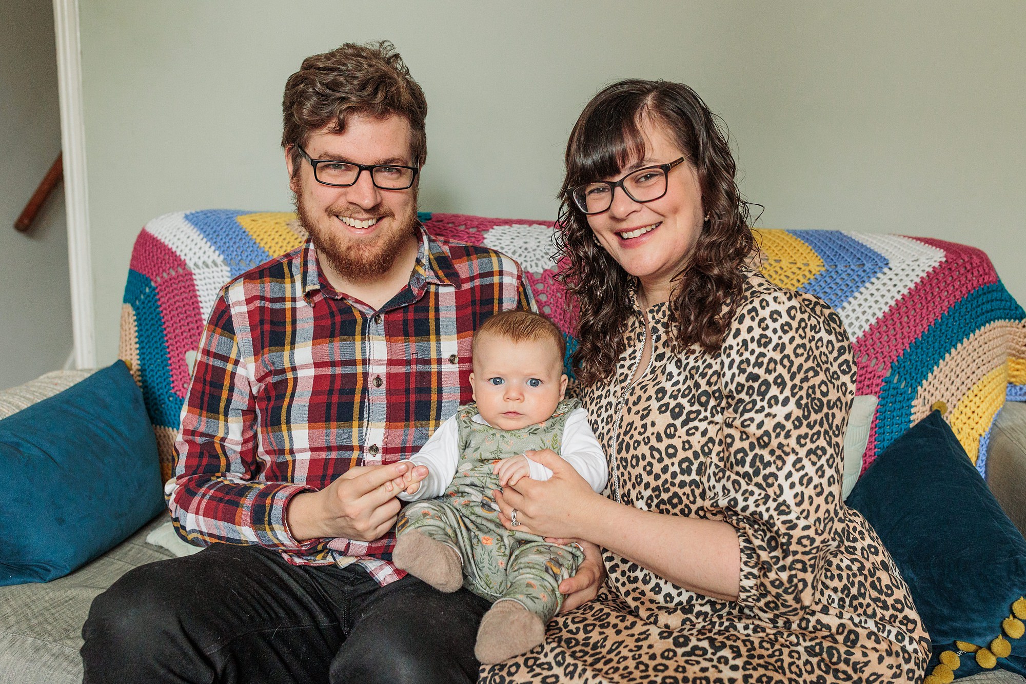 A man and a woman sit on a couch holding a baby between them. The man wears a plaid shirt, and the woman wears a leopard print dress. A colorful crocheted blanket is draped over the back of the couch.
