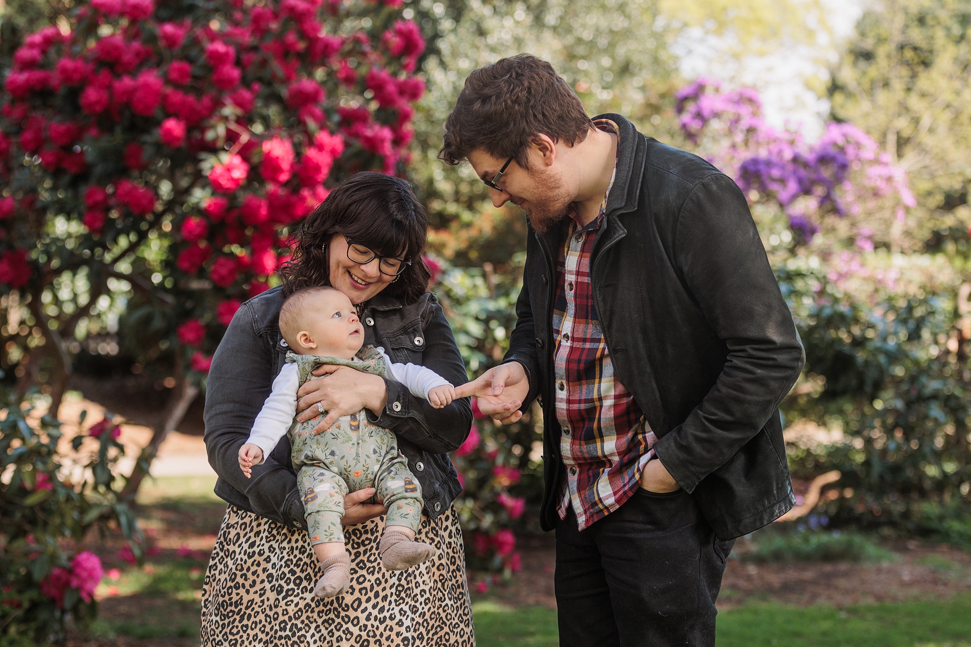 A woman holding a baby and a man standing beside them are outside in a garden with blooming red and purple flowers. The adults are smiling and looking at the baby. The baby looks up at the woman.