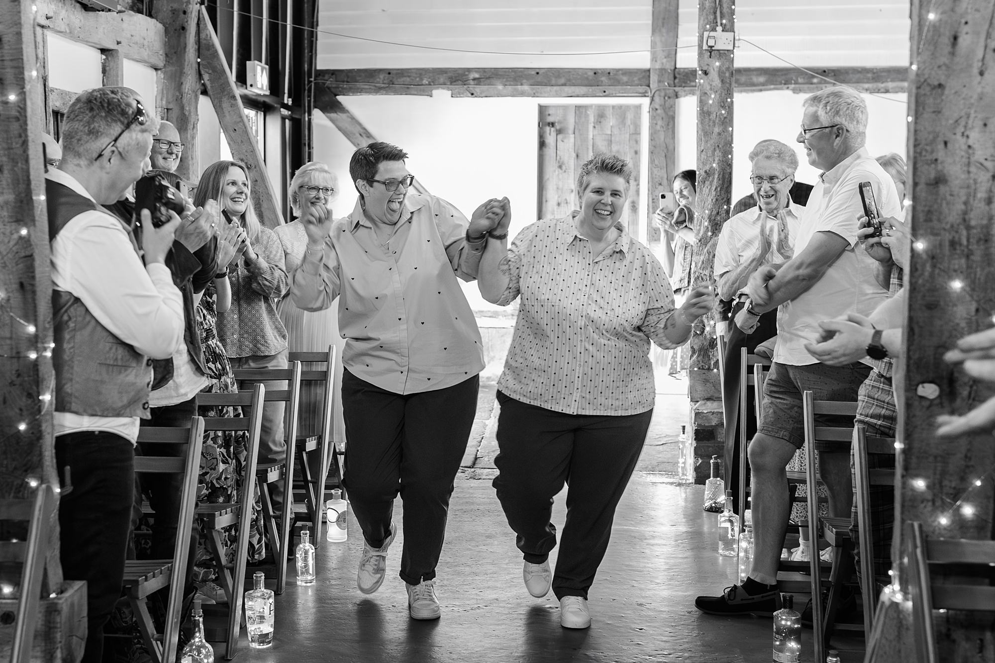 Two people walk hand-in-hand down an aisle, smiling, while guests on either side clap and take photos. The setting appears to be indoors, possibly a wedding or celebration, with string lights visible.