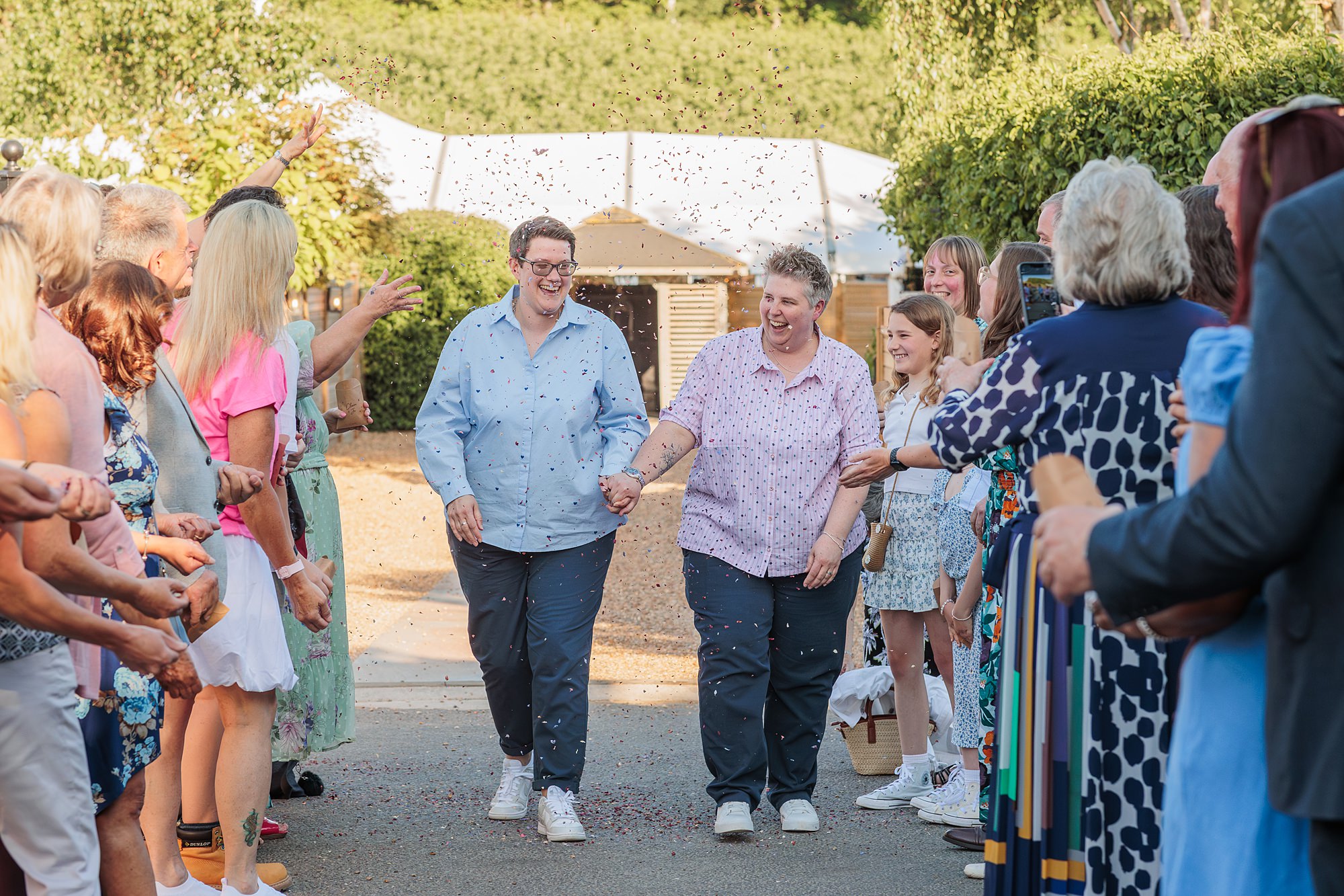 Two people holding hands walk down an outdoor aisle while guests on both sides throw confetti. Both are smiling and dressed in button-down shirts and sneakers. The scene appears to be part of a celebration or ceremony.