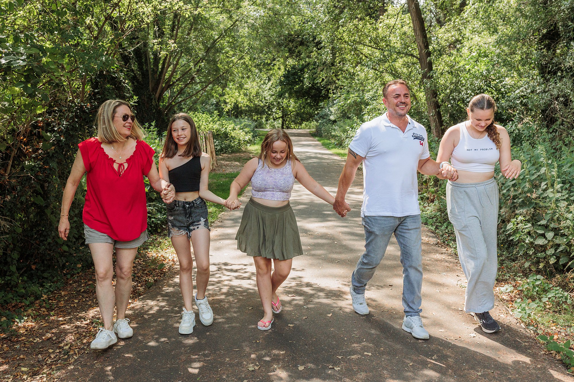 A group of five people, two adults and three children, walk hand in hand down a tree-lined path on a sunny day. They are smiling and casually dressed.