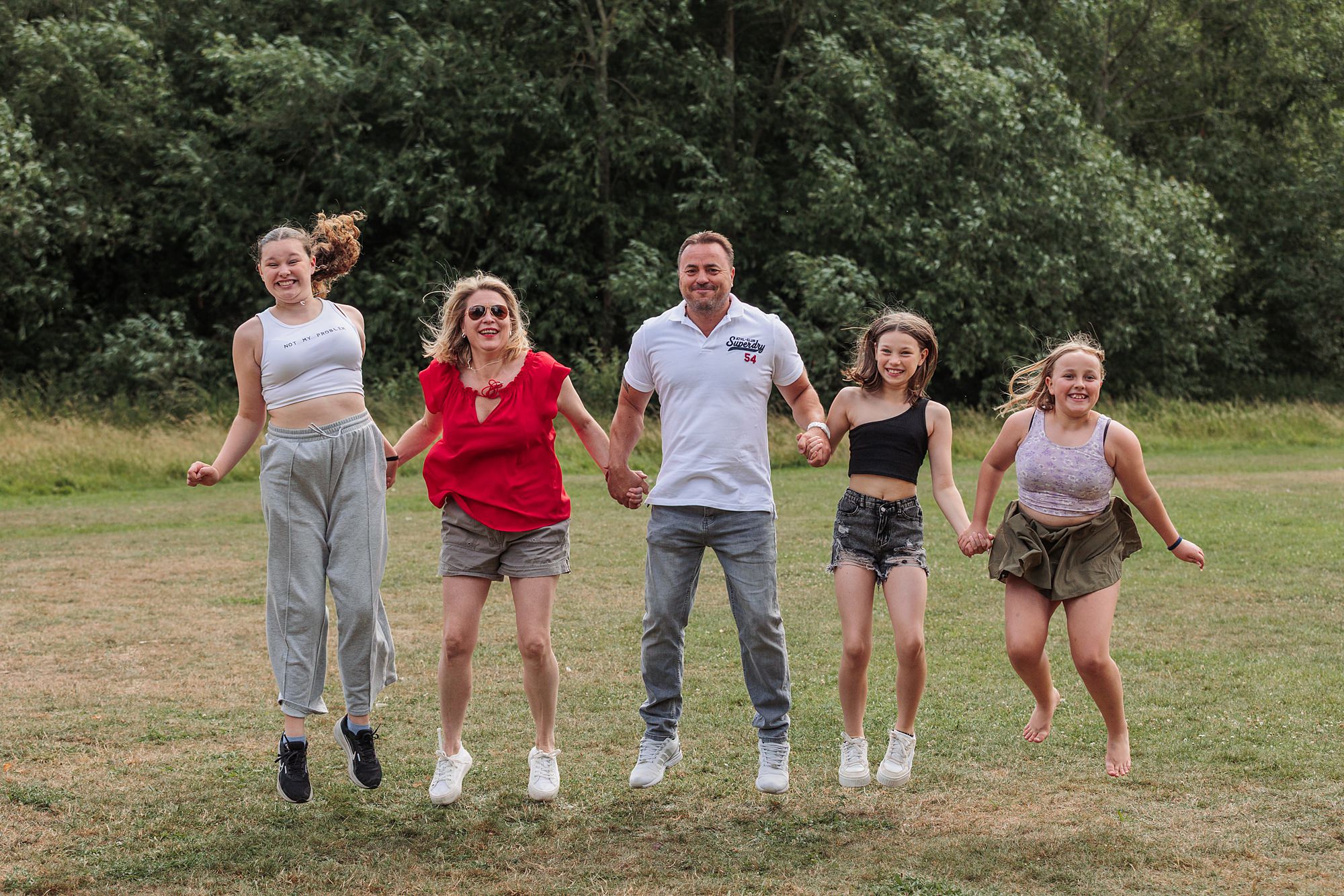 Five people, four females and one male, are holding hands and jumping together outdoors on a grassy field with trees in the background. They are smiling and wearing casual summer clothes.