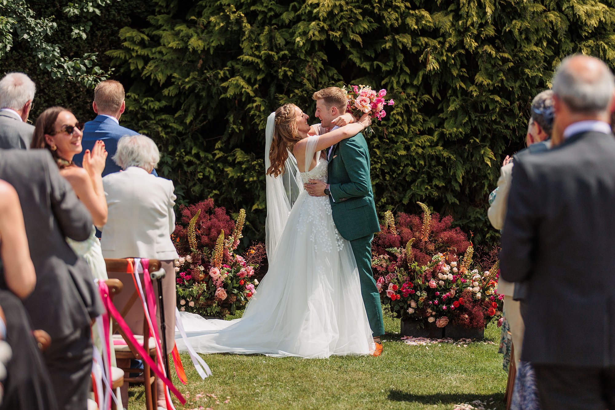 A bride and groom kiss at an outdoor wedding ceremony, surrounded by guests standing and clapping. The couple stands in front of floral arrangements and greenery on a sunny day.