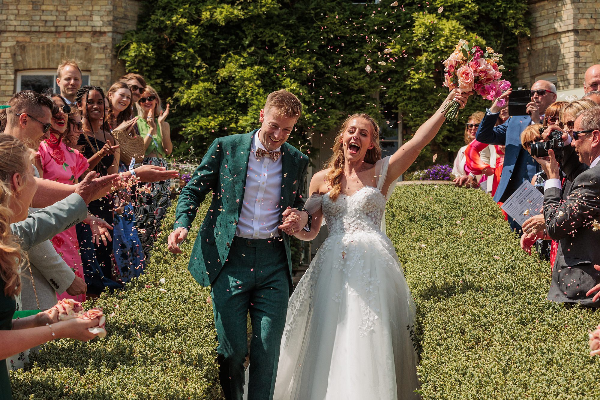 A bride and groom walk outdoors through a cheering crowd, with the bride holding a bouquet and raising her arm. Guests throw confetti, and several people are taking photos. Bushes and a stone building are in the background.