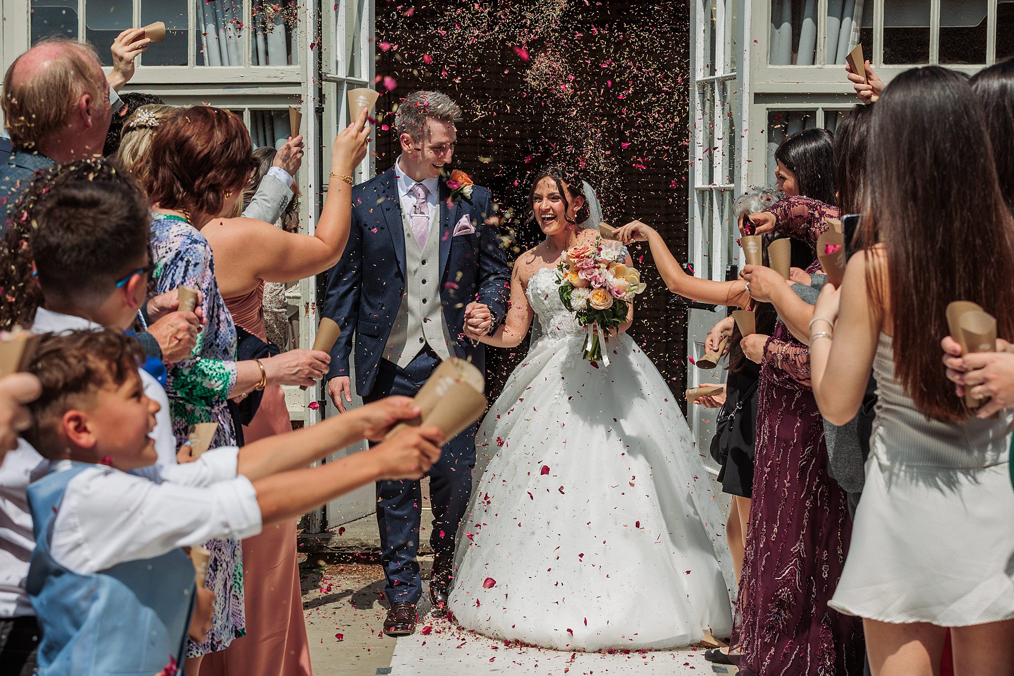 A bride and groom walk outdoors as guests throw confetti. The couple smiles and hold hands, with the bride carrying a bouquet. People stand on both sides of the path, celebrating the newlyweds in daylight.