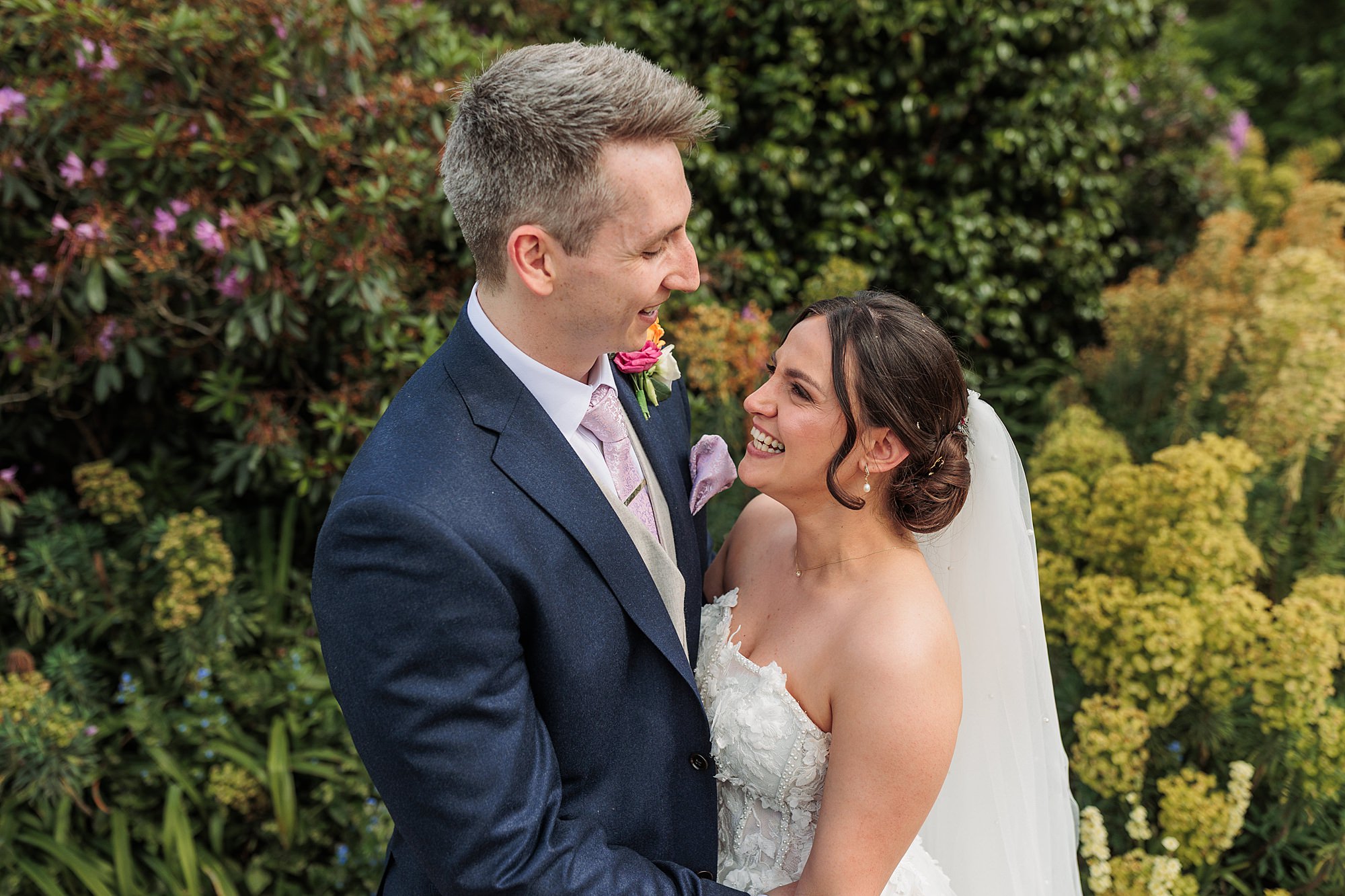 A bride in a white dress and veil smiles at a groom in a navy suit and pink tie. They stand close together outdoors, surrounded by green plants and flowers.