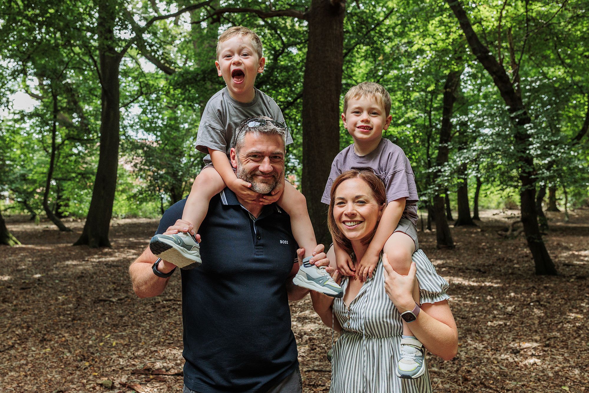 A man and woman stand in a wooded area, each carrying a young boy on their shoulders. All four are smiling or laughing, and the ground is covered with dry leaves. Sunlight filters through the trees.