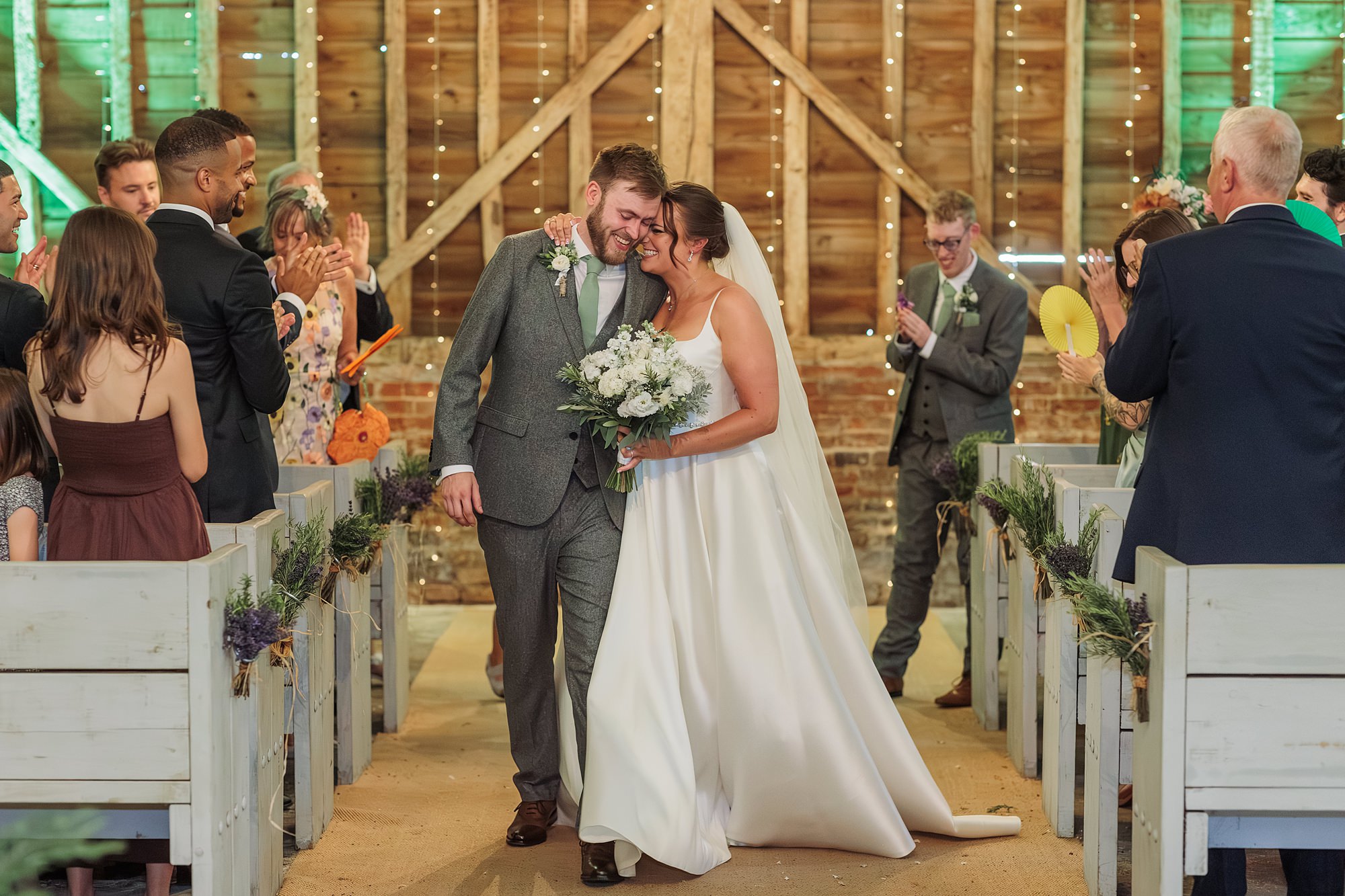 A bride and groom walk down the aisle smiling, surrounded by clapping guests in a barn decorated with string lights and rustic wooden panels. The bride holds a bouquet and wears a white dress and veil.