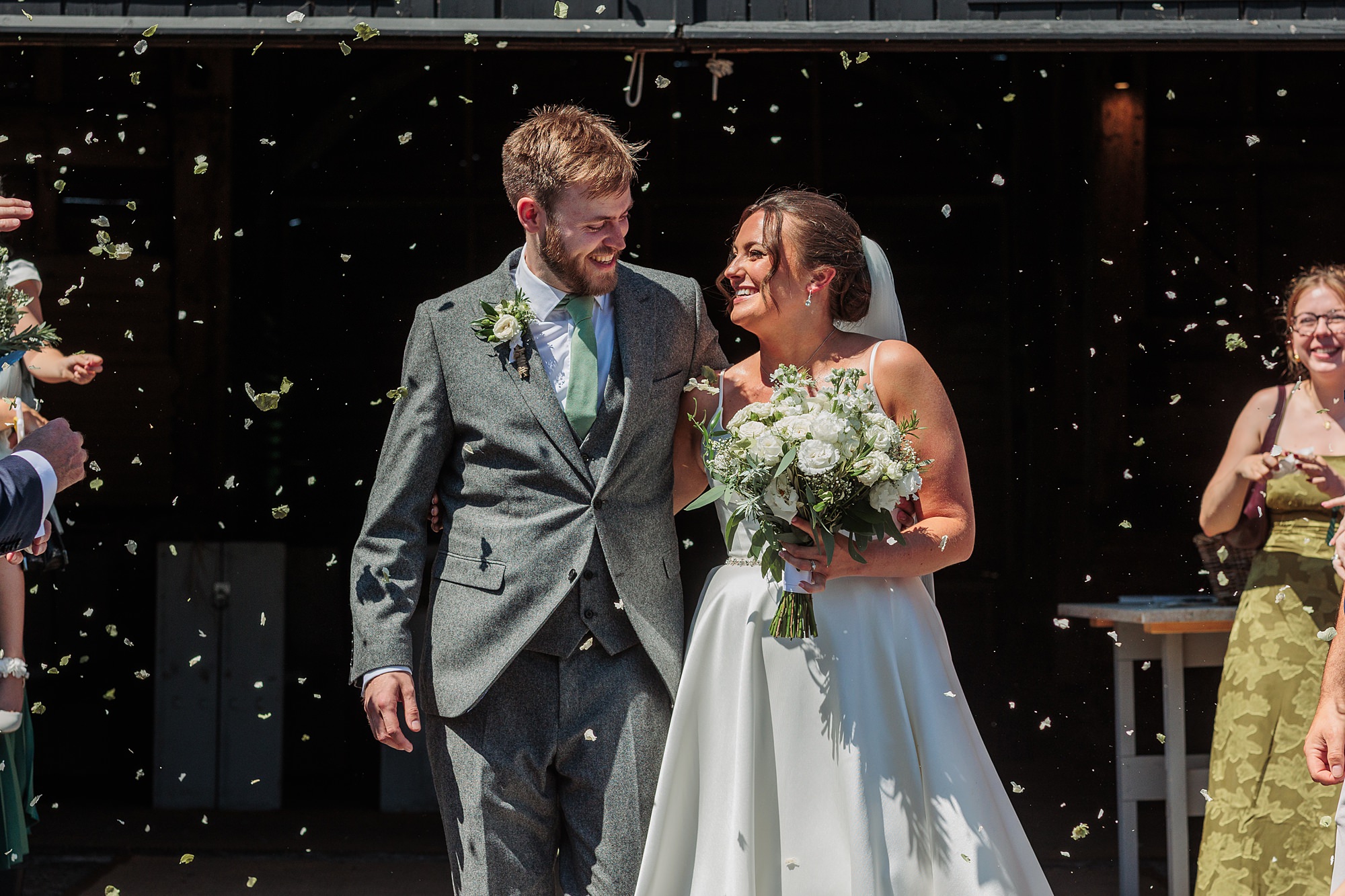 A bride and groom walk arm in arm, smiling at each other, surrounded by guests throwing confetti. The bride holds a bouquet of white flowers and wears a white dress and veil. The groom is dressed in a grey suit and green tie.