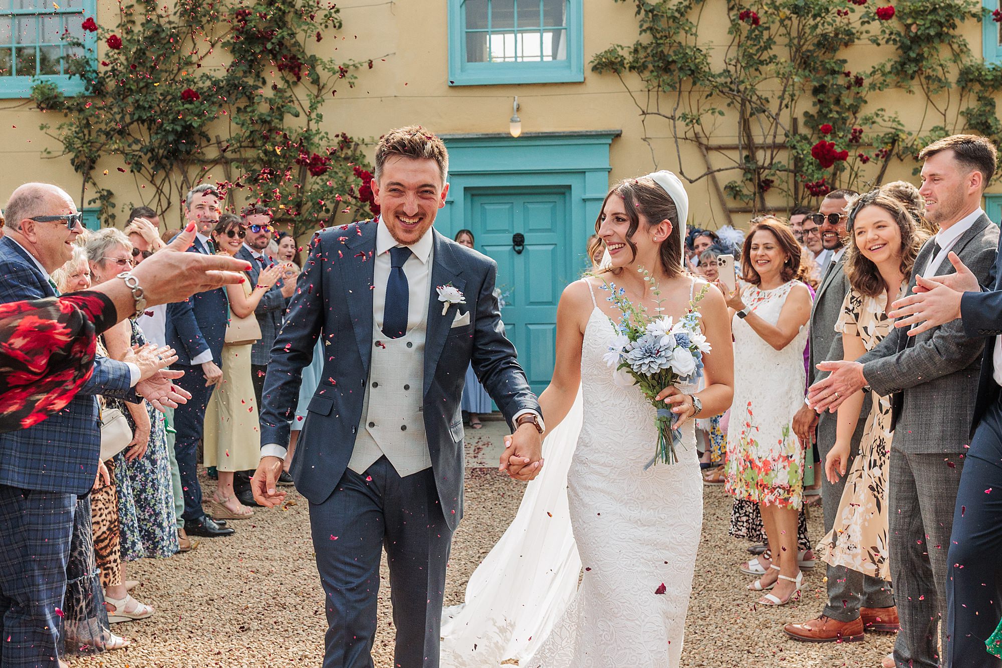 A bride and groom smile and hold hands while walking outside among guests who are throwing confetti. The groom wears a blue suit and the bride wears a white dress and holds a bouquet. The event takes place in front of a yellow building.
