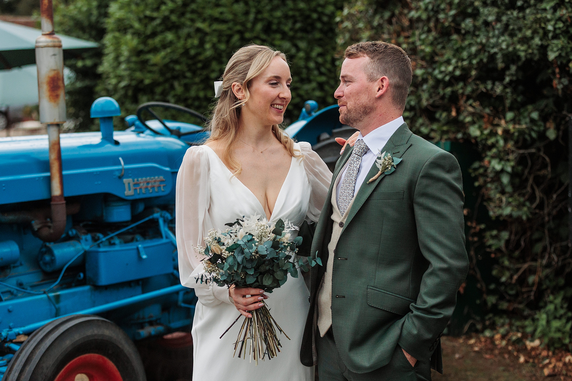 A bride in a white dress holding a bouquet stands beside a groom in a green suit. They are looking at each other and smiling, with a blue tractor and green foliage in the background.