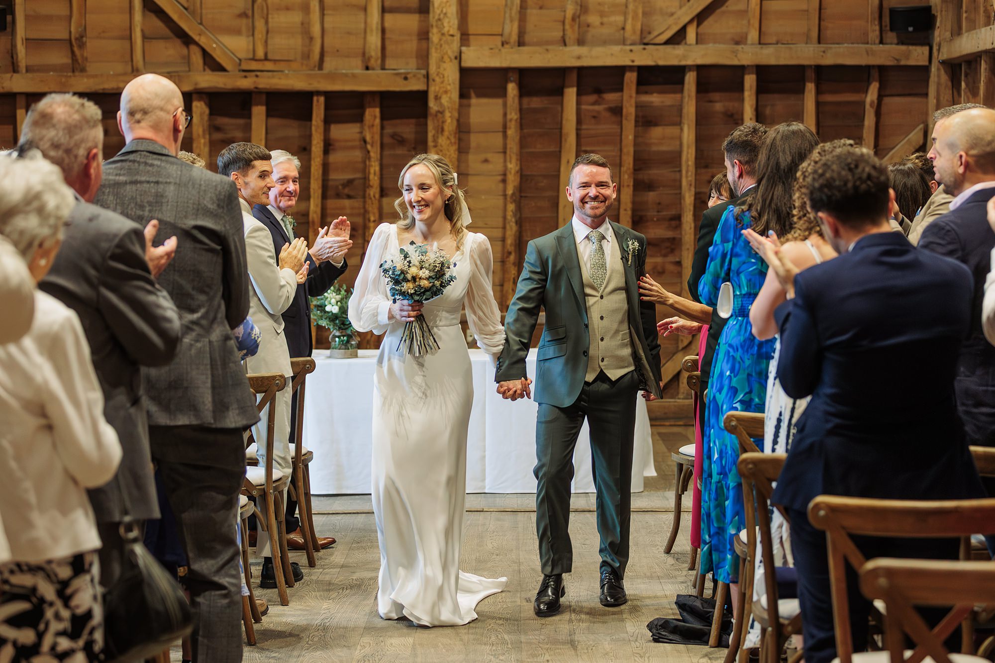 A bride and groom walk down the aisle holding hands in a rustic wooden venue. Guests seated on both sides stand and clap as the couple smiles, with the bride holding a bouquet of flowers.